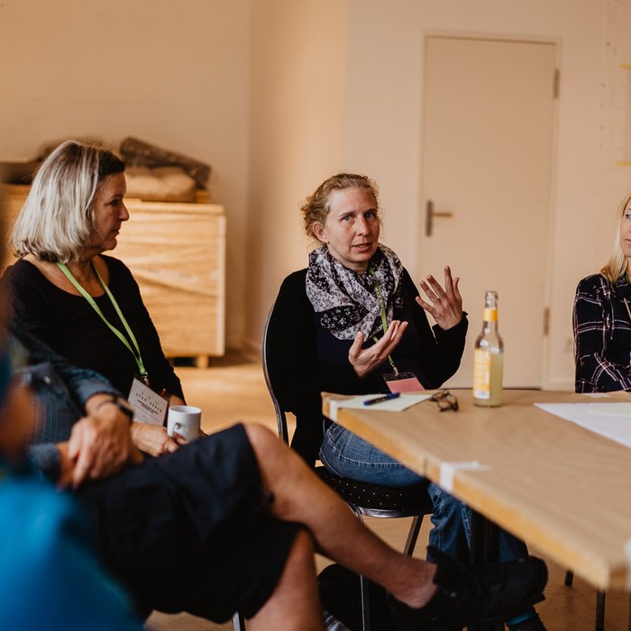 Im Vordergrund ist ein Tisch zu sehen, um den mehrere Personen sitzen. Fünf sind im Bild zu sehen. Die zweite Person von rechts spricht gerade und gestikuliert dazu. Ihre Sitznachbarinnen schauen sie an. Auf dem Tisch liegen Moderationsmaterialien.