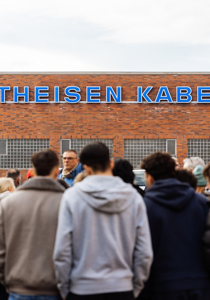 Die Menschen des Workshops stehen im Vordergrund vor der Schranke und im Hintergrund ist die Vorderwand der Kabelfabrik und der Schriftzug "Theisen Kabel" zu sehen