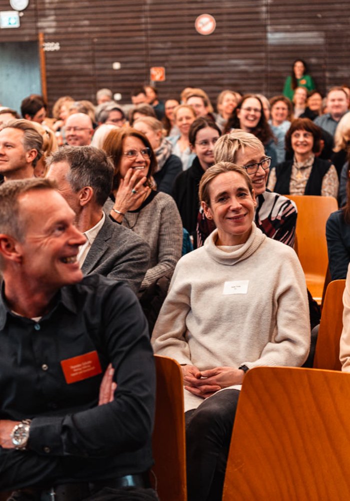 Netzwerktreffen Ganztag und Raum der Montag Stiftung Jugend und Gesellschaft: Auf Stuhlreihen sitzen sehr viele Menschen, offenbar guter Laune, ein Mann im Vordergrund in der Unschärfe dreht sich halb zu den anderen um, als habe hinten jemand etwas erheiterndes gesagt 