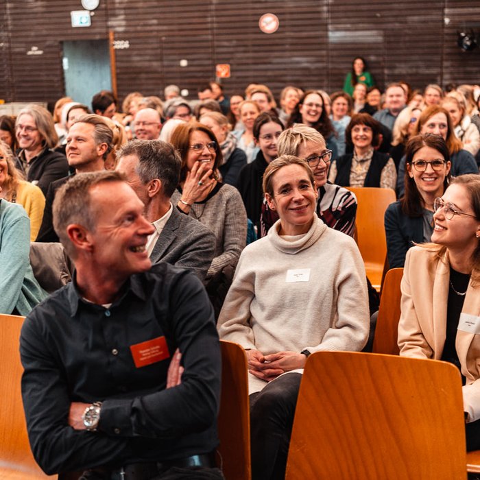 Netzwerktreffen Ganztag und Raum der Montag Stiftung Jugend und Gesellschaft: Auf Stuhlreihen sitzen sehr viele Menschen, offenbar guter Laune, ein Mann im Vordergrund in der Unschärfe dreht sich halb zu den anderen um, als habe hinten jemand etwas erheiterndes gesagt 