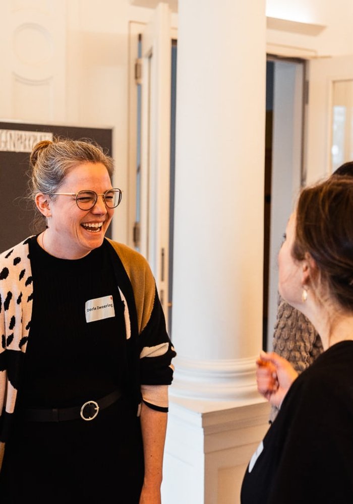 Netzwerktreffen Ganztag und Raum der Montag Stiftung Jugend und Gesellschaft: Zwei Frauen stehen sich gegenüber und reden miteinander, die eine ist von der Kamera abgewandt, auf dem Gesicht der anderen ist ein herzhaftes Lachen zu sehen; dahinter eine Stellwand mit Post-its