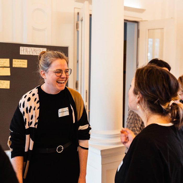 Netzwerktreffen Ganztag und Raum der Montag Stiftung Jugend und Gesellschaft: Zwei Frauen stehen sich gegenüber und reden miteinander, die eine ist von der Kamera abgewandt, auf dem Gesicht der anderen ist ein herzhaftes Lachen zu sehen; dahinter eine Stellwand mit Post-its