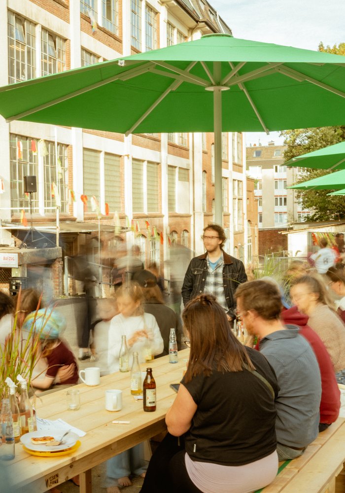Viele Menschen sitzen an langen Tischen unter grünen Schirmen