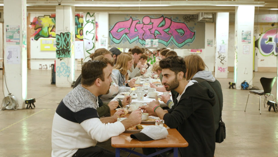 Eine Gruppe von Menschen sitzt an einem langen Tisch und isst. Man guckt vom Kopf des Tischs auf zwei Männer im Vordergrund, die am Ende des Tischs sitzen.